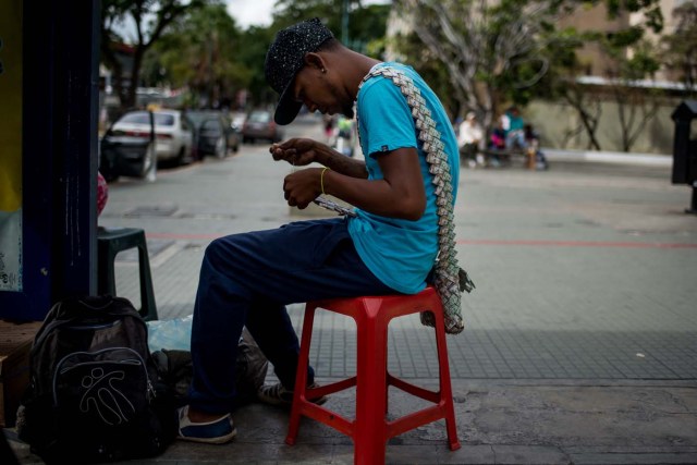 Wilmer Rojas, 25, sists at a bus stop in Caracas as he sews Bolivar bills, to make crafts on January 30, 2018. A young Venezuelan tries to make a living out of devalued Bolivar banknotes by making crafts with them. / AFP PHOTO / FEDERICO PARRA